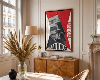 Op de afbeelding: Een ingelijste Bauhaus-poster met een rood en zwart geometrisch ontwerp hangt boven een houten kast. De poster toont een zwart-wit afbeelding van een gebouw met de tekst "BAUHAUS" en de data "1919-1928". Een vaas met gedroogde planten staat op een tafel.