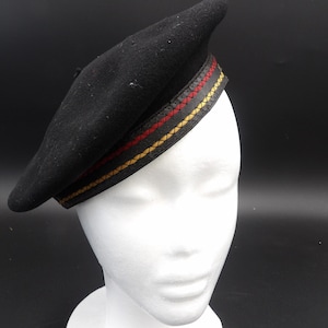 May include: Black beret with a decorative band featuring red and yellow stitching. The beret is displayed on a white mannequin head against a dark background. A classic hat style.
