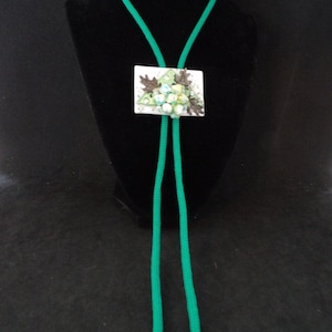 Könnte beinhalten: Eine grüne Bolo-Krawatte mit einem rechteckigen Anhänger mit floralem Design. Der Anhänger hat einen weißen Hintergrund mit einem Cluster aus hellblauen und grünen Blumen und braunen Blättern. Die Krawatte wird auf einer schwarzen Schaufensterpuppe präsentiert.