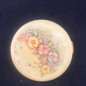 May include: A round, off-white compact with a floral design. The compact features a cluster of pansies in shades of pink, yellow, and blue, with green leaves and small pink flowers. The background has a soft, watercolor effect.
