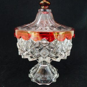May include: A clear, cut-glass candy dish with a lid, featuring a red and gold rim. The dish has a pedestal base and a decorative finial on top. The glass is cut in a diamond pattern, adding to its vintage charm.