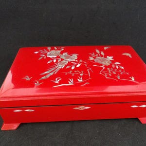 May include: A rectangular red jewelry box with a glossy finish. The lid is adorned with a floral and bird design inlaid with mother-of-pearl. The box sits on four small feet.