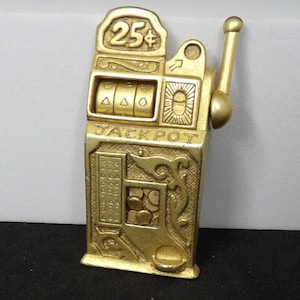 May include: A gold-toned metal slot machine figurine featuring a lever on the right side, three spinning reels, and the text 25 cents at the top with the word JACKPOT embossed across the center. The front displays a coin slot and decorative details.
