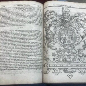 May include: Open book with aged, yellowed pages. The left page features dense text, while the right page displays a detailed black and white illustration of a coat of arms with the words "DIEV ET MON DROITS" at the bottom.