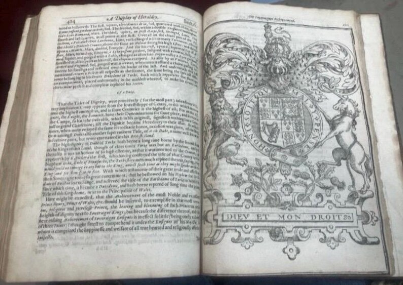 May include: Open book with aged, yellowed pages. The left page features dense text, while the right page displays a detailed black and white illustration of a coat of arms with the words "DIEV ET MON DROITS" at the bottom.
