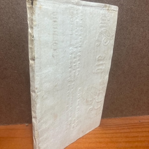 May include: A rectangular, off-white book with a textured, embossed cover featuring faint, stylized lettering and decorative floral patterns. The spine is worn with visible creases, and the edges of the cover show signs of age and slight discoloration.