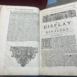 May include: Open book with aged, off-white pages. The text reads "A Display of Heraldry" with ornate decorative elements. The book is open to reveal detailed text and illustrations, showcasing a historical document.