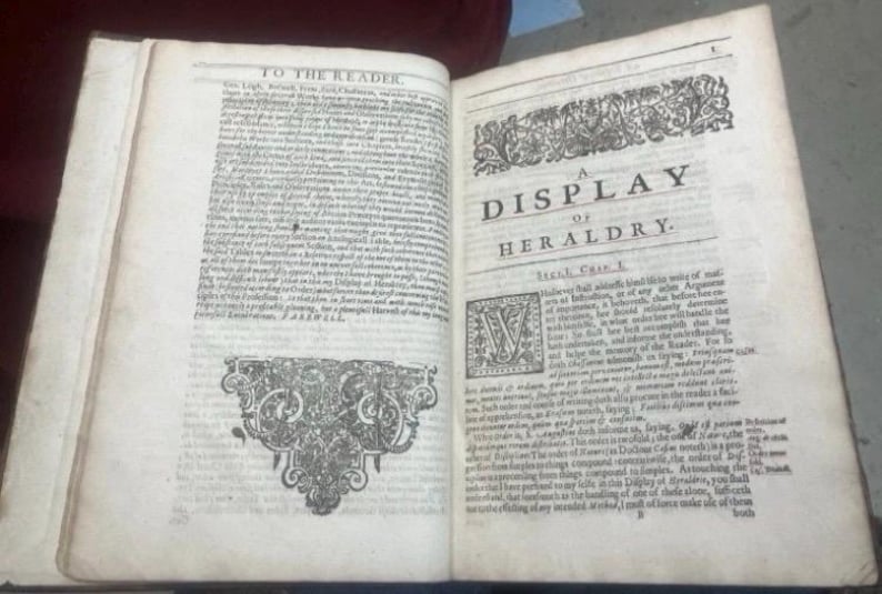 May include: Open book with aged, off-white pages. The text reads "A Display of Heraldry" with ornate decorative elements. The book is open to reveal detailed text and illustrations, showcasing a historical document.