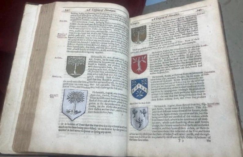 May include: Open pages of an antique book with aged, yellowed paper. The text is in old English, with decorative tree emblems. The book is open to display the text and illustrations.