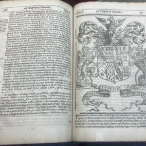 May include: An open antique book with aged, yellowed pages. The left page is filled with small text, while the right page features a detailed black and white illustration of a coat of arms with the words "DEFENDE LE DROIT" on a banner.