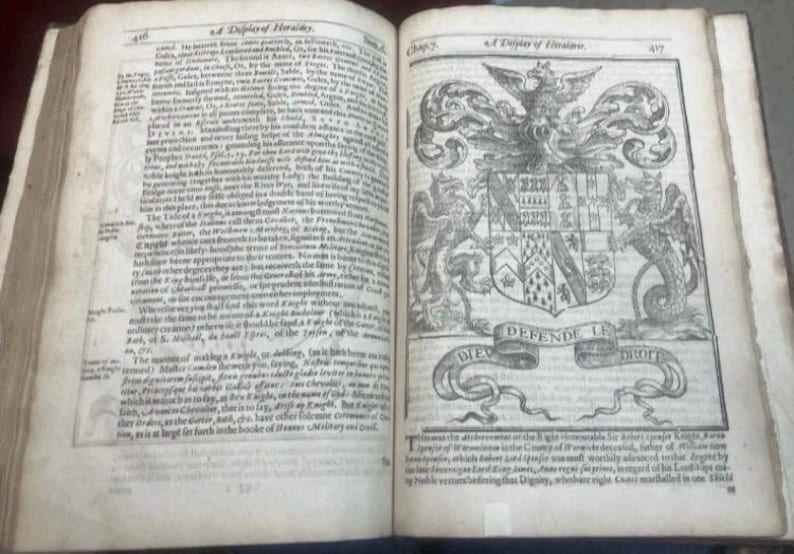 May include: An open antique book with aged, yellowed pages. The left page is filled with small text, while the right page features a detailed black and white illustration of a coat of arms with the words "DEFENDE LE DROIT" on a banner.