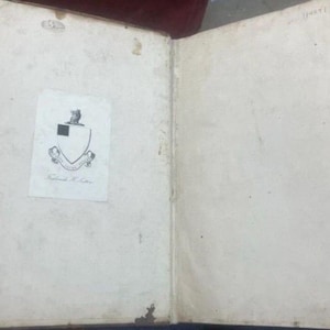 May include: Open antique book with aged, off-white pages. A small, printed paper with a crest and text is affixed to the left page. The book's binding is visible, and the pages show signs of age and wear.