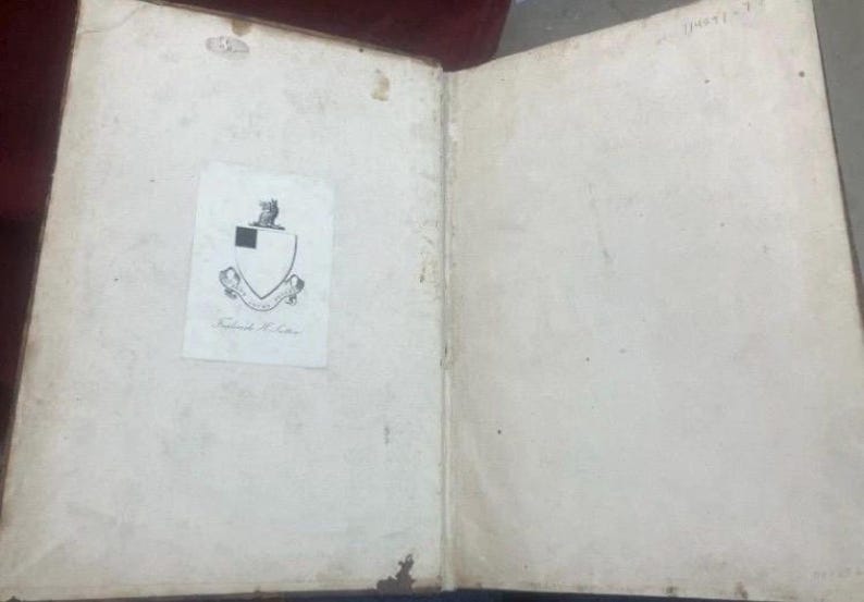 May include: Open antique book with aged, off-white pages. A small, printed paper with a crest and text is affixed to the left page. The book's binding is visible, and the pages show signs of age and wear.