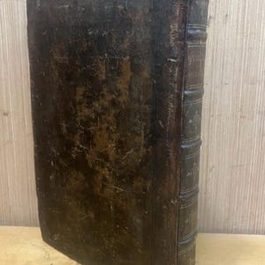 May include: An antique book with a worn, dark brown leather cover. The spine features raised bands and faded text. The edges of the pages are visible, suggesting age and use. The book is positioned on a wooden surface.