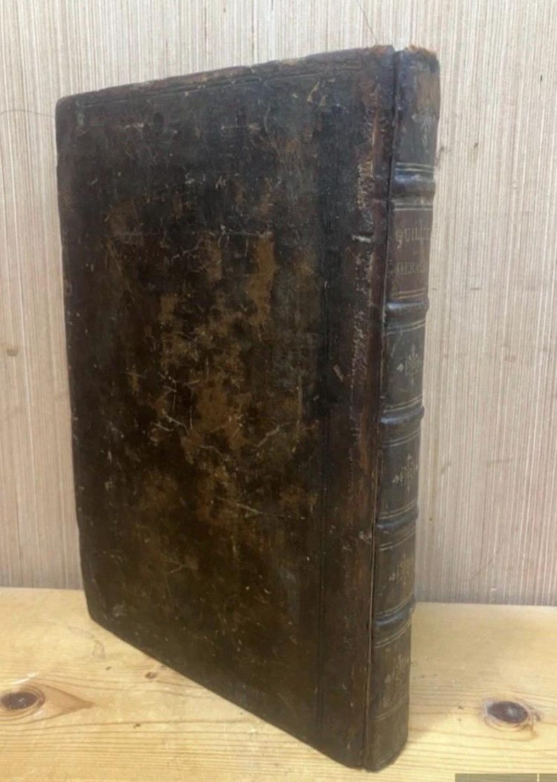May include: An antique book with a worn, dark brown leather cover. The spine features raised bands and faded text. The edges of the pages are visible, suggesting age and use. The book is positioned on a wooden surface.