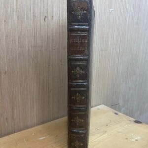 May include: A vintage, leather-bound book with the title "Guillim's Heraldry" embossed in gold lettering. The spine is dark brown with decorative gold accents and raised bands. The book appears aged, with wear and tear visible on the cover.