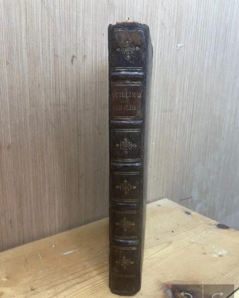 May include: A vintage, leather-bound book with the title "Guillim's Heraldry" embossed in gold lettering. The spine is dark brown with decorative gold accents and raised bands. The book appears aged, with wear and tear visible on the cover.