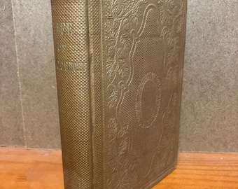 May include: A vintage hardcover book with a textured gold-toned cover featuring an embossed floral pattern and a central oval frame. The spine shows a grid-like texture and faint, partially obscured lettering near the top edge.
