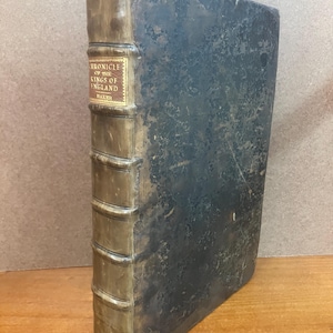 May include: Antique book titled "Chronicle of the Kings of England" by Baker. The book has a dark, weathered cover and a tan spine with gold lettering. The edges of the pages are visible, and the book appears to be well-worn.
