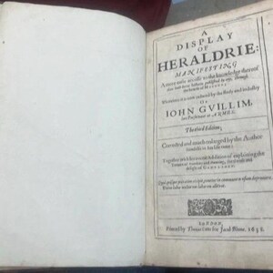 May include: An open book with aged, off-white pages. The right page features a title page with the text "A DISPLAY OF HERALDRIE: MANIFESTING". The book is an antique, likely a historical reference.