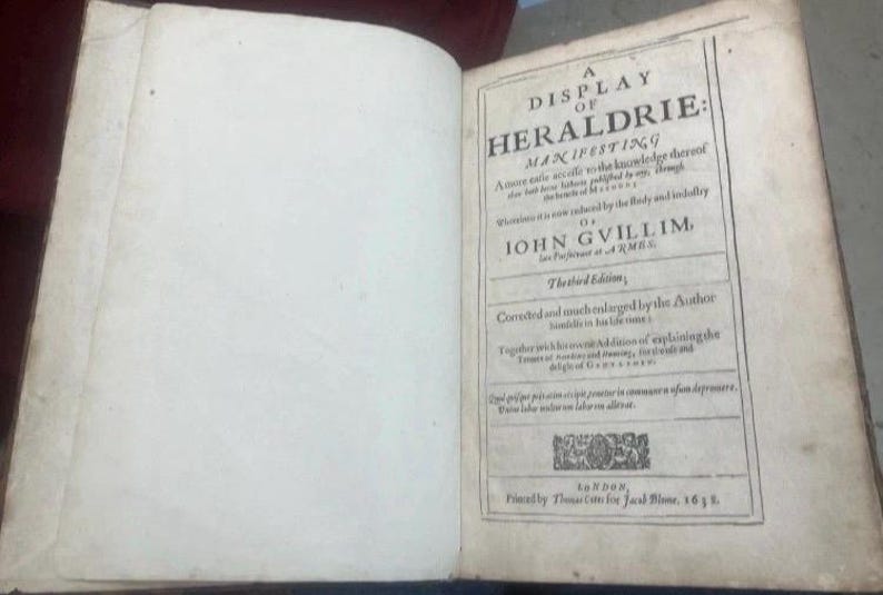 May include: An open book with aged, off-white pages. The right page features a title page with the text "A DISPLAY OF HERALDRIE: MANIFESTING". The book is an antique, likely a historical reference.