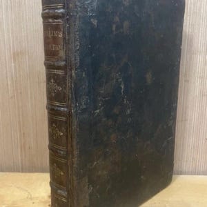 May include: An antique book with a dark leather cover and spine. The spine is brown with gold lettering and decorative accents. The book's pages are aged, and the overall appearance suggests it is a vintage item.