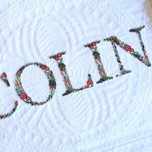 May include: A white quilted blanket featuring the name COLIN embroidered in large capital letters. Each letter is intricately filled with a colourful botanical pattern of green leaves, small red mushrooms, and various tiny floral elements.