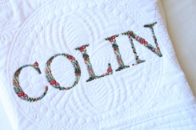 May include: A white quilted blanket featuring the name COLIN embroidered in large capital letters. Each letter is intricately filled with a colourful botanical pattern of green leaves, small red mushrooms, and various tiny floral elements.