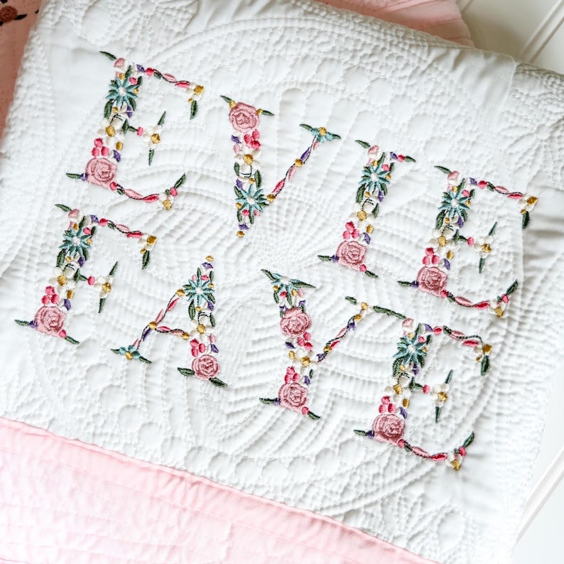 May include: A white quilted blanket featuring the embroidered name Evie Faye in a floral font. The letters are composed of colourful pink roses, green leaves, and small yellow accents, set against a textured white fabric with a circular pattern.