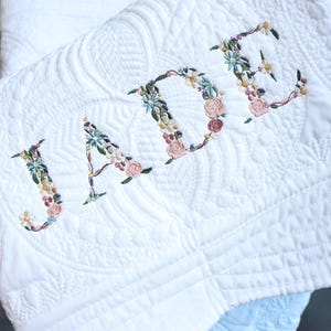May include: A white quilted blanket featuring the name JADE embroidered in a floral pattern. The letters are composed of small, colourful stitched flowers in shades of pink, blue, yellow, and green, set against a textured, stitched white fabric.