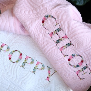 May include: A white quilted blanket featuring the name Poppy in floral appliqu&eacute; letters and a pink quilted blanket featuring the name Chloe in floral appliqu&eacute; letters, both with intricate stitched patterns across the fabric surfaces.