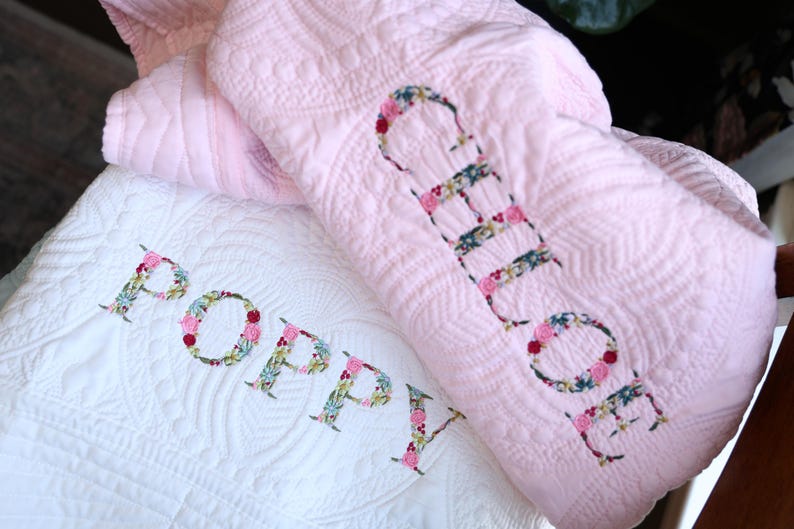 May include: A white quilted blanket featuring the name Poppy in floral appliqu&eacute; letters and a pink quilted blanket featuring the name Chloe in floral appliqu&eacute; letters, both with intricate stitched patterns across the fabric surfaces.