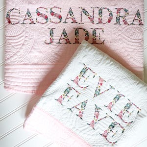 May include: A light pink quilted blanket embroidered with the name CASSANDRA JADE and a white quilted blanket embroidered with the name EVIE FAYE. Both blankets feature floral patterned lettering in shades of pink, green, and dark blue.