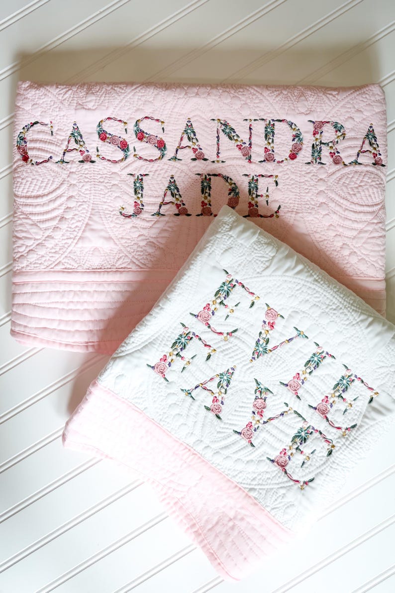 May include: A light pink quilted blanket embroidered with the name CASSANDRA JADE and a white quilted blanket embroidered with the name EVIE FAYE. Both blankets feature floral patterned lettering in shades of pink, green, and dark blue.