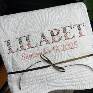 May include: A white quilted blanket featuring the embroidered name LILABET in a floral pattern and the date September 17, 2025, in pink thread. The folded fabric is tied with a thin olive green ribbon centred across the front surface.