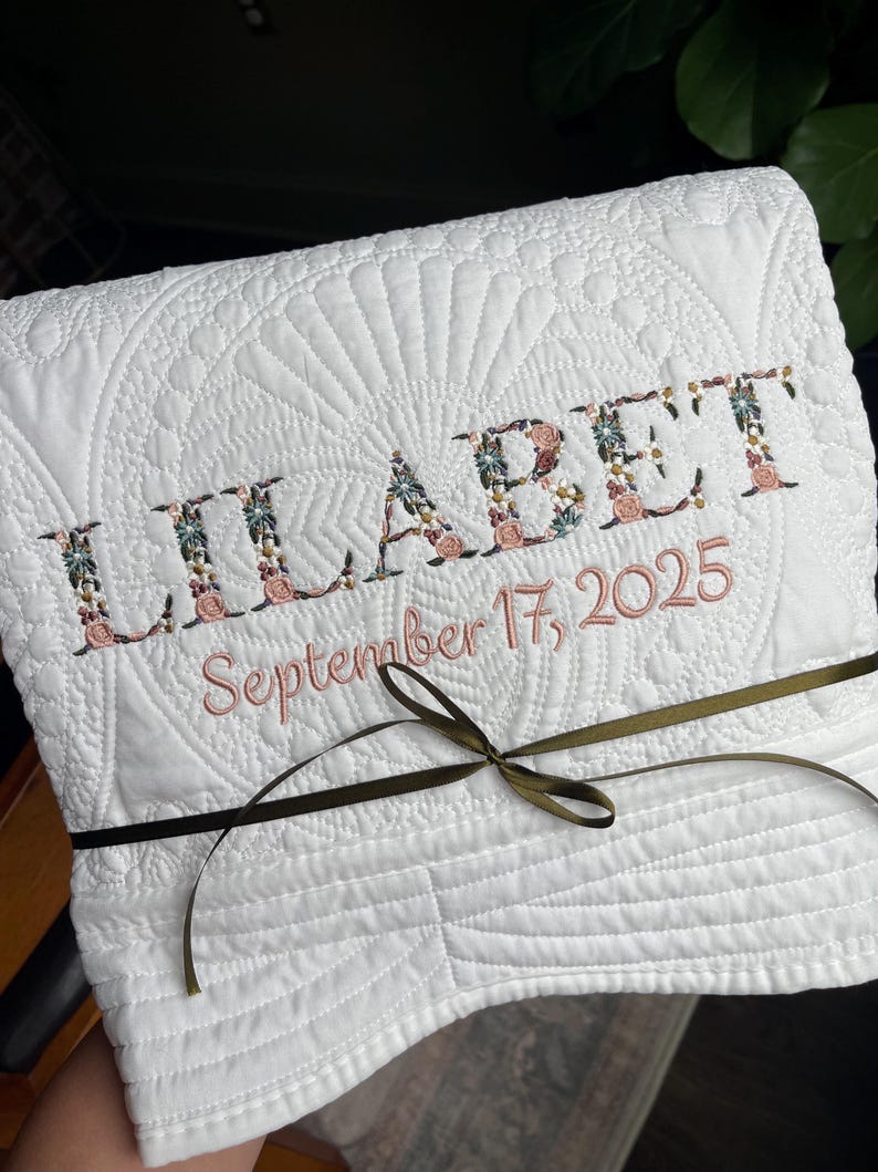 May include: A white quilted blanket featuring the embroidered name LILABET in a floral pattern and the date September 17, 2025, in pink thread. The folded fabric is tied with a thin olive green ribbon centred across the front surface.