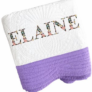 May include: A white quilted blanket featuring the name ELAINE embroidered in a floral pattern. The blanket has a solid lavender-coloured section at the bottom with horizontal stitching and a white top section with a decorative quilted fan design.