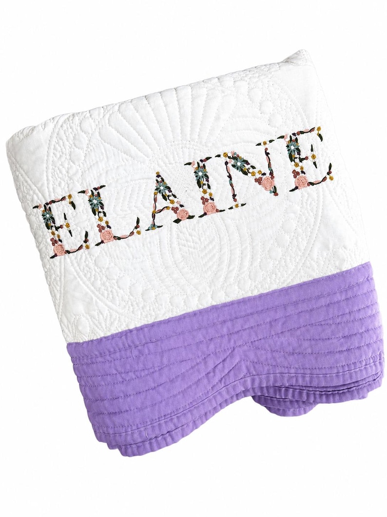 May include: A white quilted blanket featuring the name ELAINE embroidered in a floral pattern. The blanket has a solid lavender-coloured section at the bottom with horizontal stitching and a white top section with a decorative quilted fan design.