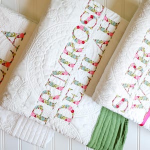 May include: Three white quilted blankets featuring intricate stitched patterns. Each blanket is personalised with large, floral-patterned fabric letters spelling out the names ELLA, PRIMROSE DELILAH, and SABRINA WILSON, with coloured fabric borders.