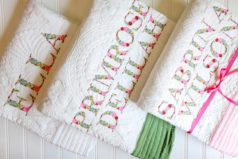 May include: Three white quilted blankets featuring intricate stitched patterns. Each blanket is personalised with large, floral-patterned fabric letters spelling out the names ELLA, PRIMROSE DELILAH, and SABRINA WILSON, with coloured fabric borders.