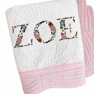 May include: A white quilted blanket featuring the name ZOE embroidered in a floral pattern with pink, green, and brown tones. The blanket has a solid light pink border along the bottom edge and a textured, stitched design throughout the white fabric.