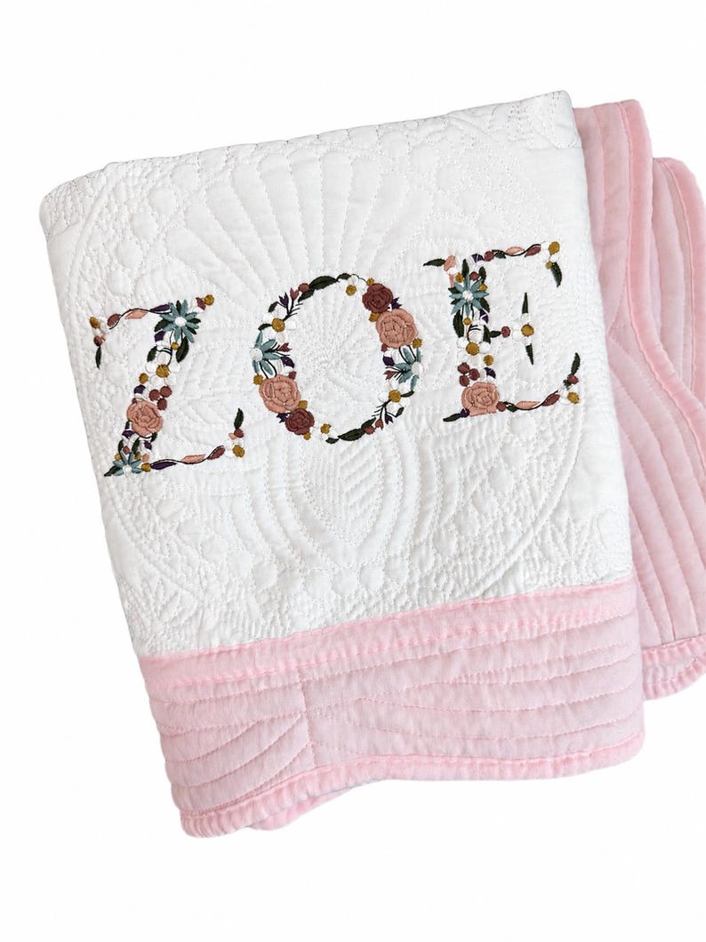 May include: A white quilted blanket featuring the name ZOE embroidered in a floral pattern with pink, green, and brown tones. The blanket has a solid light pink border along the bottom edge and a textured, stitched design throughout the white fabric.