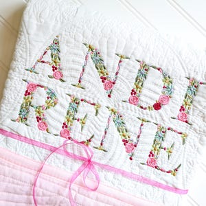 May include: White quilted baby blanket embroidered with the name "ANDI RENE" in floral letters. The letters are filled with green leaves, red berries, and pink roses. A pink ribbon is tied in a bow at the bottom.