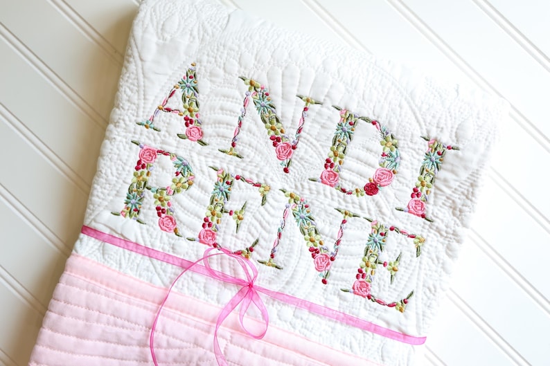 May include: A white quilted blanket featuring the embroidered name ANDI REINE in floral-patterned letters. The quilt has a textured, stitched surface and a light pink horizontal band near the bottom, accented with a thin, translucent pink ribbon.