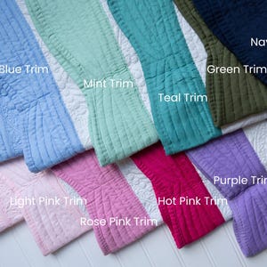 May include: A collection of quilted fabric swatches featuring white floral patterns with various coloured borders labelled Solid Blue, Blue Trim, Mint Trim, Teal Trim, Green Trim, Navy Trim, Solid Pink, Light Pink Trim, Rose Pink Trim, Hot Pink Trim, and Purple Trim.