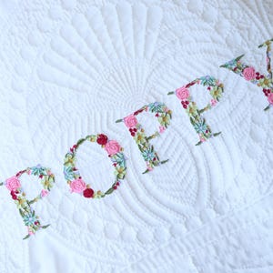 May include: A white quilted blanket featuring the name POPPY embroidered in a floral pattern. The letters are composed of small pink, red, and green thread flowers and leaves. The fabric has a subtle, stitched fan-shaped texture pattern.