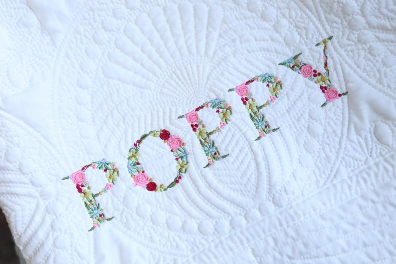 May include: A white quilted blanket featuring the name POPPY embroidered in a floral pattern. The letters are composed of small pink, red, and green thread flowers and leaves. The fabric has a subtle, stitched fan-shaped texture pattern.