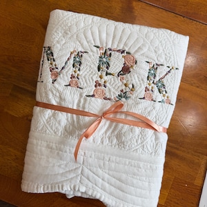 May include: A white quilted blanket featuring the embroidered letters MBK in a floral pattern. The letters are filled with small pink, green, and blue flowers. The blanket is folded and tied with a thin, peach-coloured satin ribbon across the centre.
