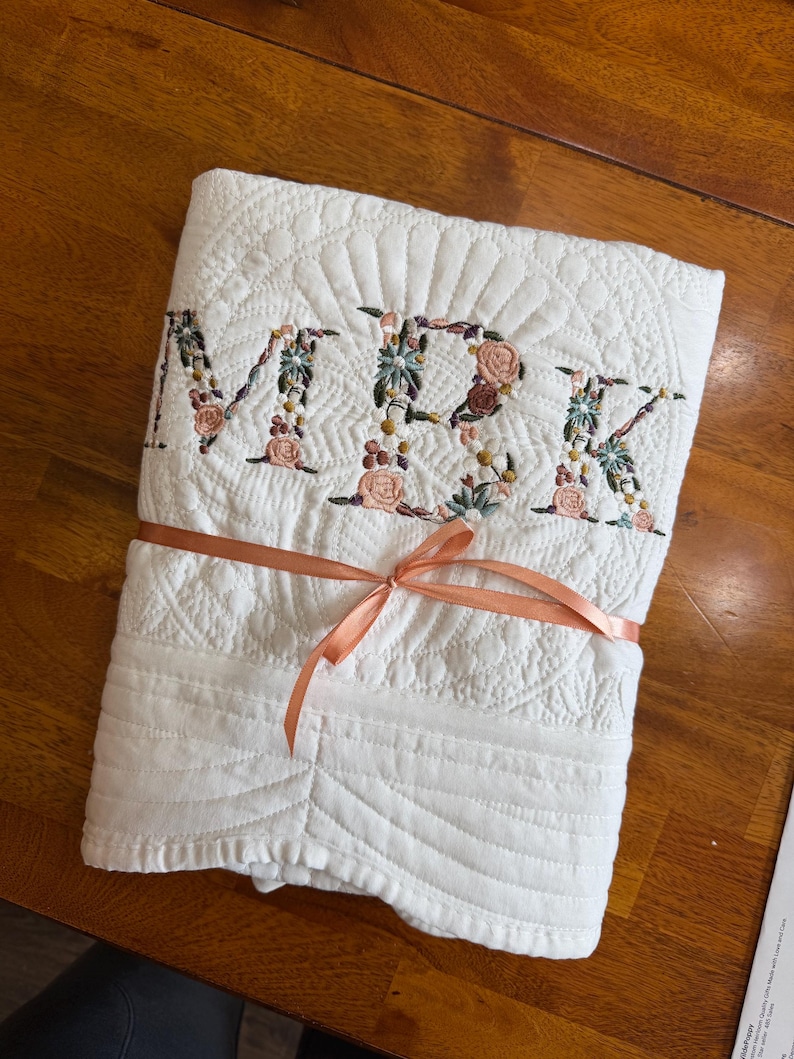 May include: A white quilted blanket featuring the embroidered letters MBK in a floral pattern. The letters are filled with small pink, green, and blue flowers. The blanket is folded and tied with a thin, peach-coloured satin ribbon across the centre.