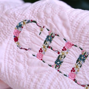 May include: A light pink quilted blanket featuring the name ELLE embroidered in a floral script. The letters are formed by intricate thread-stitched flowers in shades of pink, green, yellow, and blue, set against a textured, stitched fabric surface.